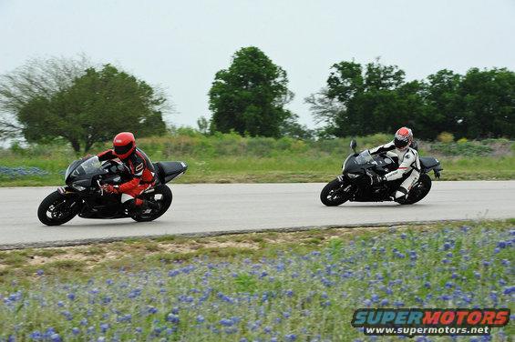 Two sport motorcycles on a racetrack, one in red gear, the other in white gear