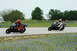 2001 Kawasaki KZ1000-A1 - Two sport motorcycles race on a track bordered by bluebonnets, with one rider in red and the other in white racing suits.