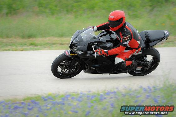 Motorcyclist in red and black gear riding a black sportbike on a paved track with blurred green grass and blue flowers in the foreground