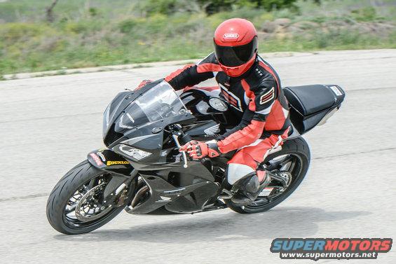 Rider in red and black gear on a black sportbike, leaning into a turn. The bike has a Honda logo visible