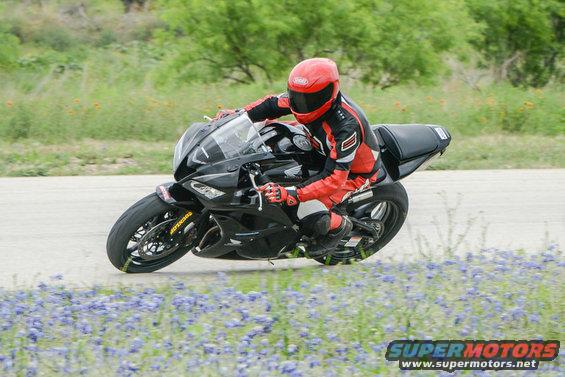 Rider in red and black gear on a black sportbike, leaning into a turn on a track with bluebonnets in the foreground