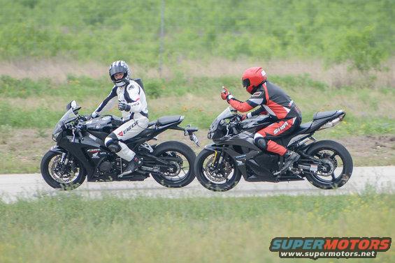 Two sport motorcycles ridden by people in full gear on a road with grassy background