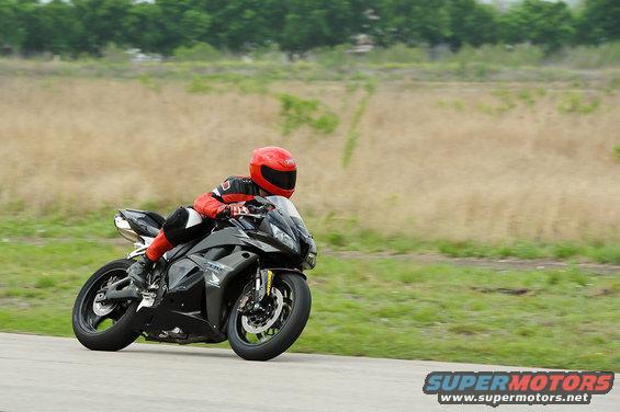 Motorcyclist in red and black gear on a black sportbike, leaning into a turn on a paved road with dry grass and trees in the background