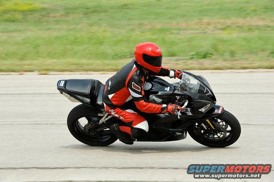 Black motorcycle side view with rider in red and black suit. Supermotors.net logo visible