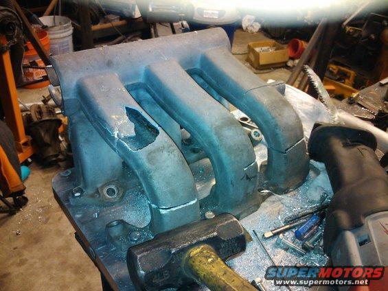 Damaged aluminum intake manifold with broken runners and tool marks, on a workbench