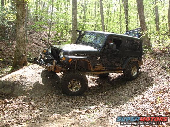 Black Jeep Wrangler Rubicon on a dirt trail, going over a rock. Another Jeep is visible behind it