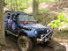 1979 Ford Bronco - A blue Jeep Wrangler equipped for off-roading navigates a wooded trail with a snorkel and roof rack visible.