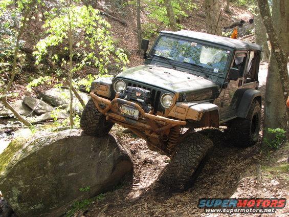 Green Jeep Wrangler climbing over a large rock on a wooded trail. It has a winch and a custom bumper