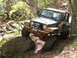 1979 Ford Bronco - A Jeep Wrangler navigates a challenging off-road obstacle on a dirt path surrounded by trees.