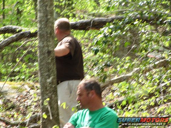 Two men are in a wooded area. One man in a brown shirt and tan pants stands behind a tree. Another man in a green shirt is in the foreground