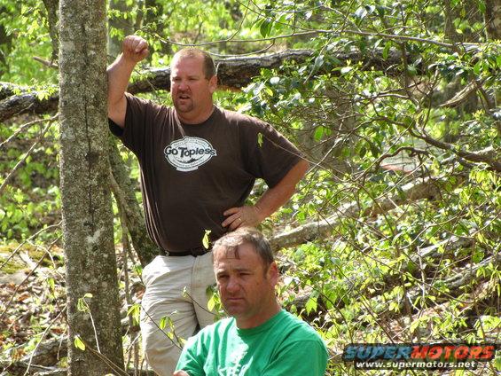 Two men stand in a wooded area, one leans against a tree. The man in front wears a green shirt, the man behind wears a brown shirt with 'Go Topless' logo