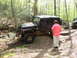 1979 Ford Bronco - A black Jeep Wrangler Rubicon is shown off-roading on a dirt trail in a wooded area, with another vehicle visible in the background.