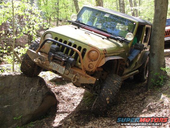 Green Jeep Wrangler climbing over a rock on a wooded trail. Another orange Jeep is visible behind it