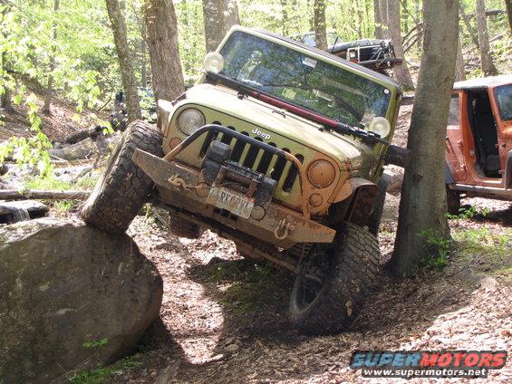 Jeep Wrangler climbing over a large rock on a wooded trail, another Jeep visible behind it