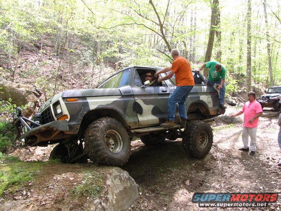 Camouflaged SUV with people climbing on it, navigating rocks on a trail. Another SUV is visible in the background