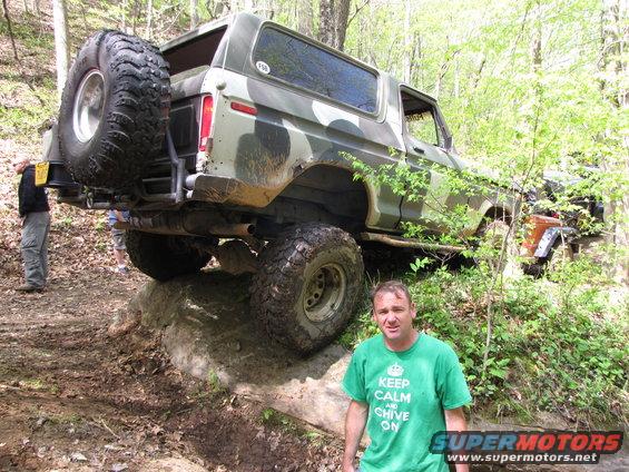Camouflaged 1978 Ford Bronco on rocky off-road trail with spare tire mounted on rear. Man in green shirt stands nearby