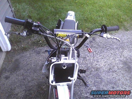 Dirt bike handlebars with "PRO TAPER" branding, black grips, and brake lever. Front number plate is visible