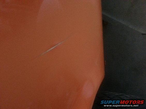 Close-up of an orange car panel showing a long scratch. A dark area and tire are visible on the right