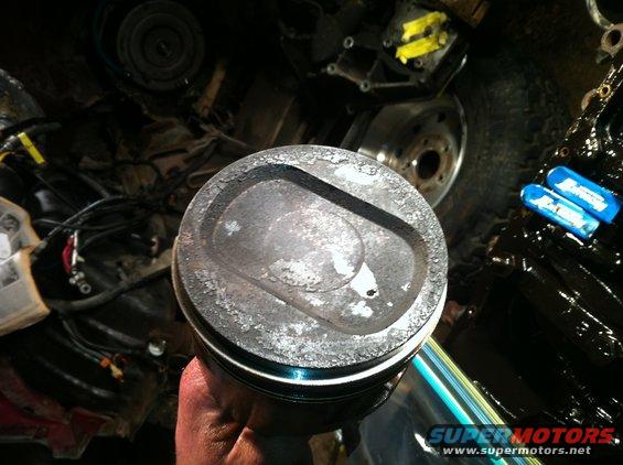 Hand holding a dirty piston with carbon buildup. Engine parts are visible in the background
