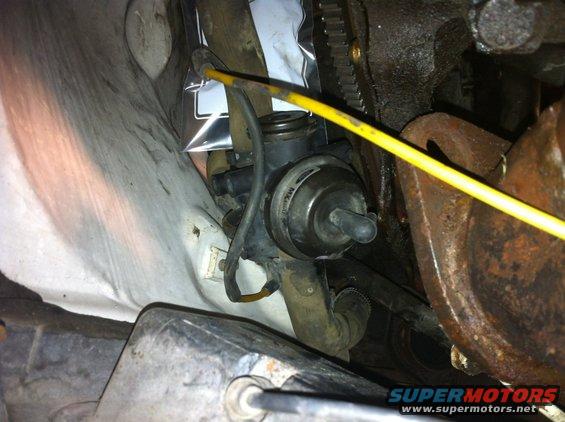 Close-up of a vacuum actuator and hoses near engine parts. A yellow rod points towards the actuator