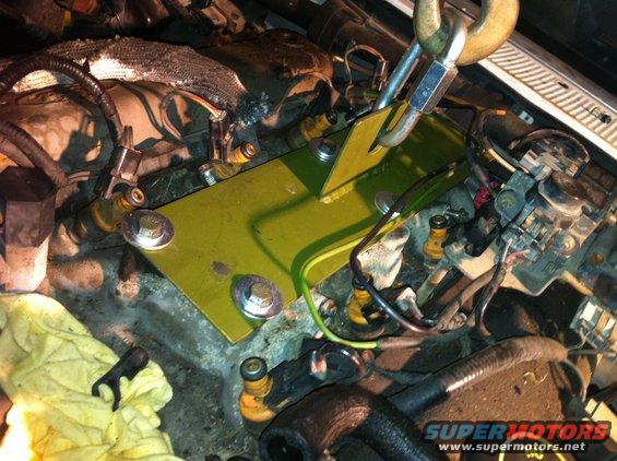 Engine bay with a yellow plate and bolts being installed, wires and hoses visible