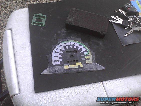 Car dashboard gauge cluster with white and green markings, keys, and a sanding block on a black surface