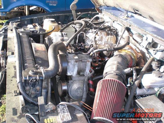 Engine bay with hoses, belts, and air filter visible. Blue truck in background