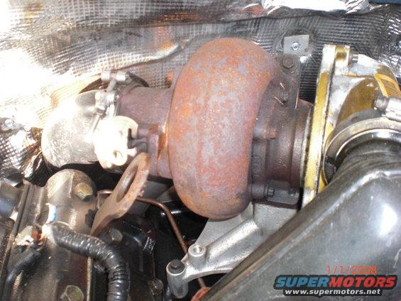 Close-up of a rusty turbocharger with heat shielding above it. Some hoses and engine parts are visible below