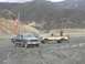 1986 Ford Bronco - Two pickup trucks are parked in a muddy area with hills in the background. An American flag flies from the blue truck.