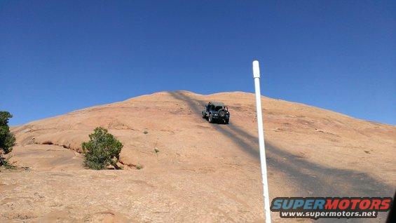 Dark SUV driving up a dirt road on a rocky hill under a clear blue sky