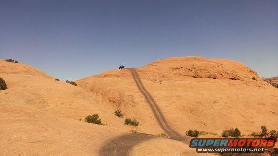 Off-road vehicle on a dirt track ascending a rocky hill under a clear blue sky