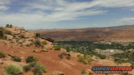 Desert landscape overlooking a town, with red rocks and sparse vegetation in the foreground. Watermark reads www.supermotors.net