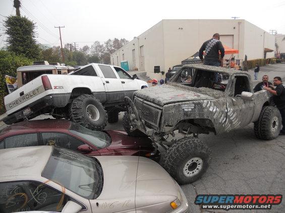 Mud-covered pickup truck with large tires, front grille visible, parked on other cars