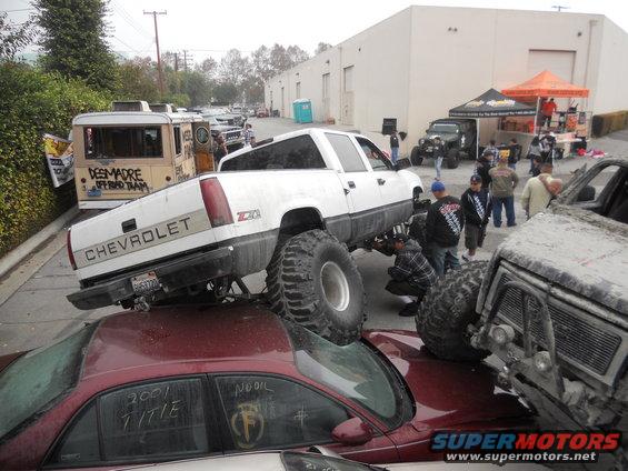 White Chevrolet pickup truck with massive tires crushing a red sedan