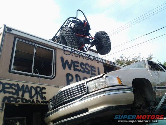 Front view of a tan RV with graffiti and a buggy on its roof, next to a tan car