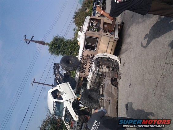 Two heavily damaged off-road trucks on pavement, one on its side. Text on one truck reads 'WESSEL GOODONE'
