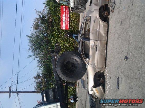 Damaged tan sedan with missing wheels, leaning on a large tire. A red sign reads "HOBBIES & TOYS"