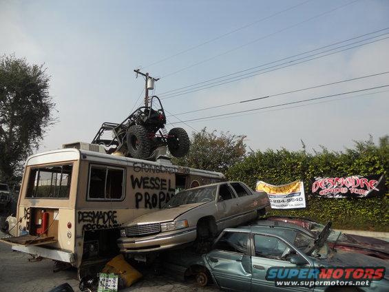 Bus with buggy on roof, stacked cars, and 'DESMADRE OFFROAD TEAM' banner
