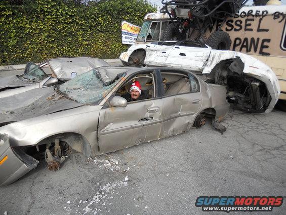 Side view of a wrecked silver sedan with a man in a Santa hat inside, other damaged vehicles behind