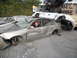 1986 Ford Bronco - A man in a Santa hat sits in a heavily damaged silver sedan, surrounded by other wrecked vehicles in a junkyard.
