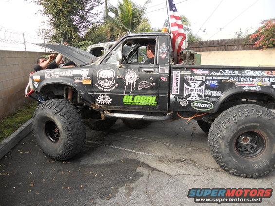 Modified Toyota pickup truck with large off-road tires, skull decals, and an American flag. Hood is open with a person working on the engine