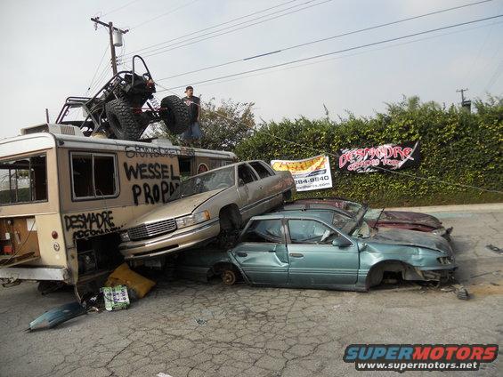 Bus with buggy on roof, gold sedan on top of teal sedan, red car behind teal sedan. Banners and graffiti on bus