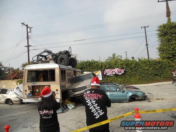 Scene of multiple wrecked vehicles, including a bus with an off-road buggy on top, and two people wearing Santa hats
