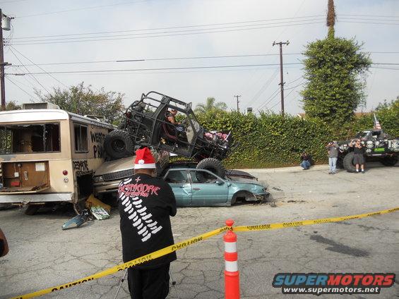 Off-road buggy atop a wrecked car and bus, with people watching. Yellow caution tape is in the foreground