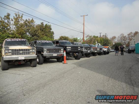 Several lifted pickup trucks and SUVs parked in a line. The first truck has a 'DESMADRE' banner on its windshield