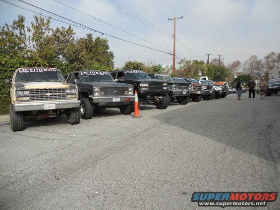 Several lifted trucks and SUVs parked in a row, some with 'DESMADRE' banners on windshields