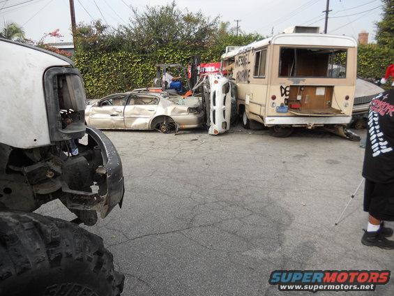 Damaged sedan and overturned vehicle next to a bus; foreground shows part of a large off-road vehicle