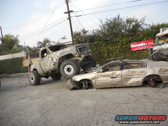 Monster truck crushing a gold sedan. Sign for 'Hobbies & Toys' in background