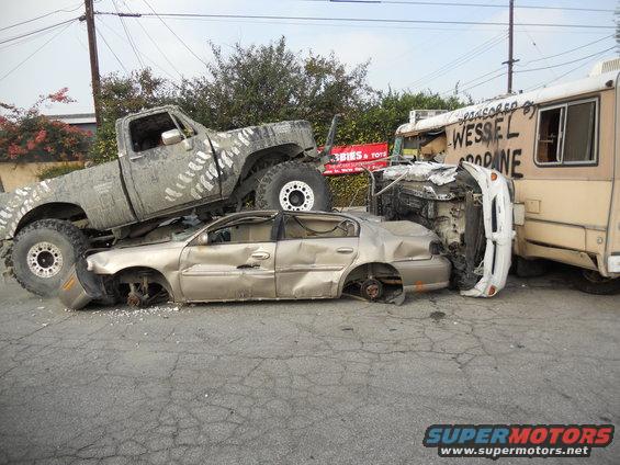 Big lifted truck on top of a wrecked sedan, next to a damaged RV