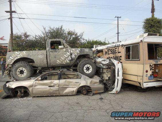 Lifted pickup truck crushing sedan, with bus behind. Text on bus reads 'WESSEL OFFROAD'